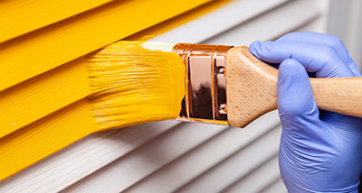 a person wearing a disposable glove using a paint brush to paint an exterior wall yellow