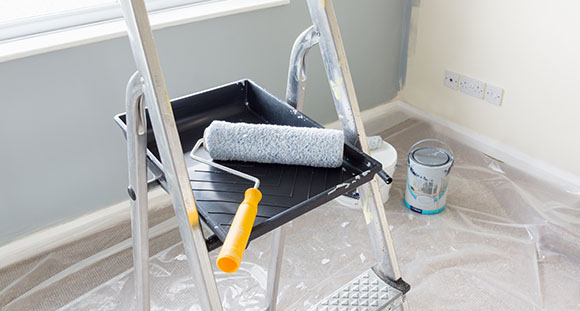 a paint roller inside a paint tray on a step ladder, in a room with a plastic sheet covering the carpet