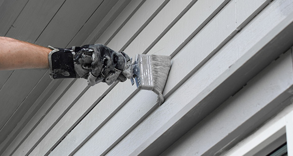 a person wearing a black paint-stained glove using a paintbrush to paint a home exterior grey